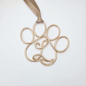 May include: A gold-colored wire paw print ornament with a tan ribbon for hanging. The paw print is made of looped wire and has a cursive "L" in the center. The ornament is on a white background.