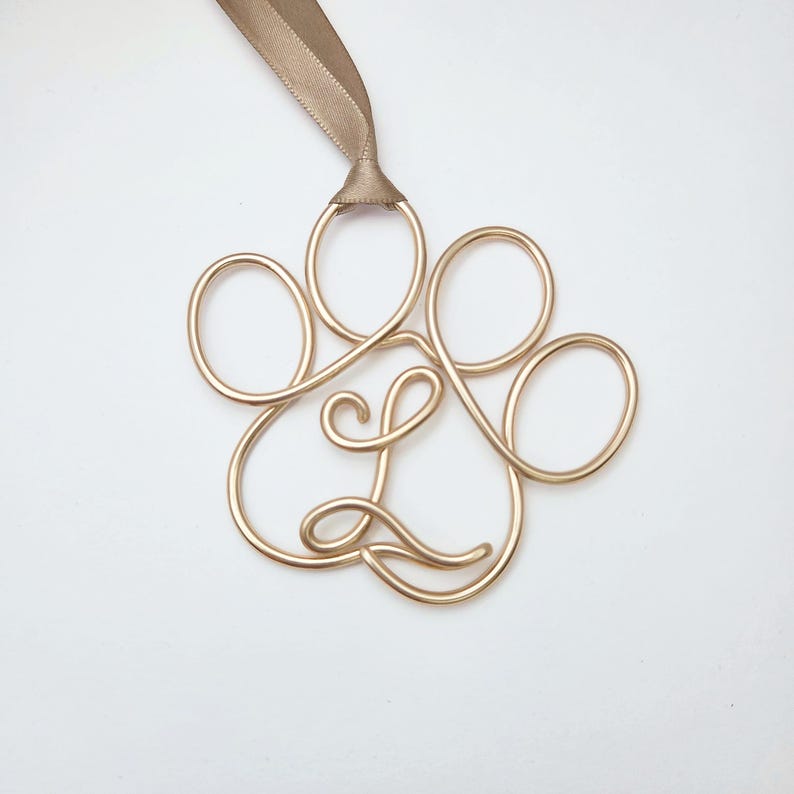 May include: A gold-colored wire paw print ornament with a tan ribbon for hanging. The paw print is made of looped wire and has a cursive "L" in the center. The ornament is on a white background.