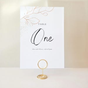 May include: A white table card with the word "One" in elegant script, accented with gold leaf design. The card is held by a gold wire holder. The text "Mia and Oliver, 11th of June" is printed at the bottom. Ideal for weddings and events.
