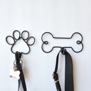 May include: Two black metal dog-shaped hooks, one shaped like a paw print and the other like a bone, are mounted on a white wall.  A black leash is hanging from each hook.