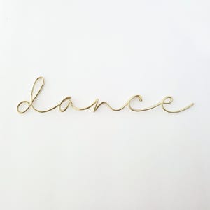 May include: A gold-colored wire sculpture spelling out the word "dance" in cursive script. The wire is bent and shaped to form each letter, creating a delicate and artistic piece against a white background. The word is centered.