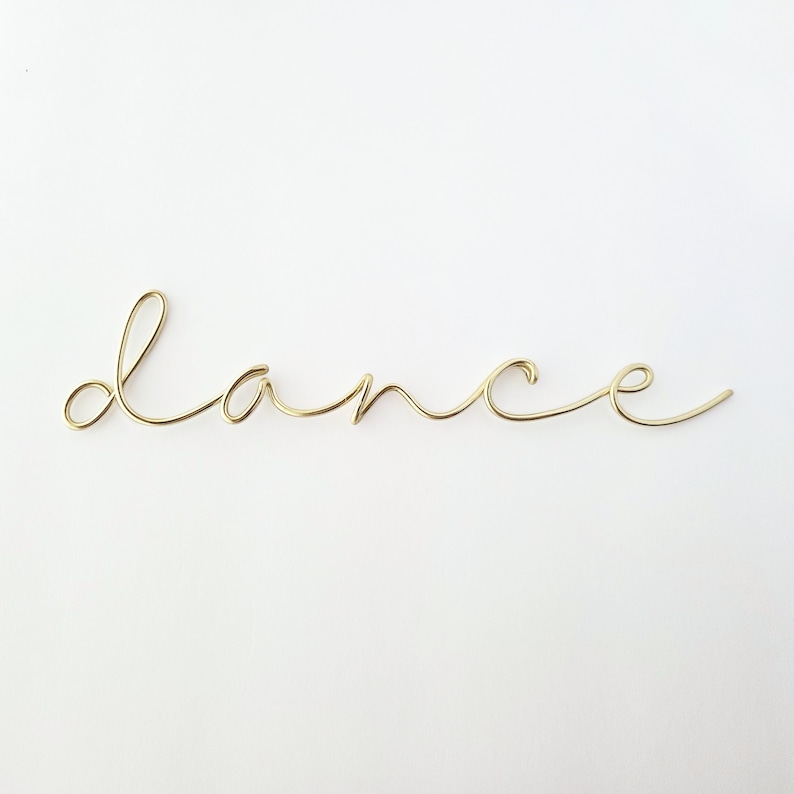 Op de afbeelding: Een goudkleurige draadsculptuur die het woord "dance" in cursief spelt. De draad is gebogen en gevormd om elke letter te vormen, waardoor een delicaat en artistiek stuk ontstaat. De sculptuur staat tegen een effen witte achtergrond.
