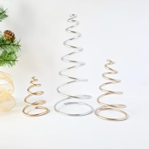 May include: Three spiral-shaped Christmas tree decorations. One is silver, and two are gold. The trees are made of metal and have a minimalist design. The trees are different sizes, with the largest being approximately 12 inches tall.