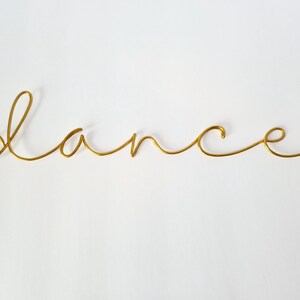 Op de afbeelding: Een goudkleurige draadsculptuur met het woord 'dance' in een cursief lettertype. Het woord is gecentreerd op een witte achtergrond. De draad is dun en heeft een gladde, glanzende afwerking, wat een minimalistische en elegante esthetiek cre&euml;ert.