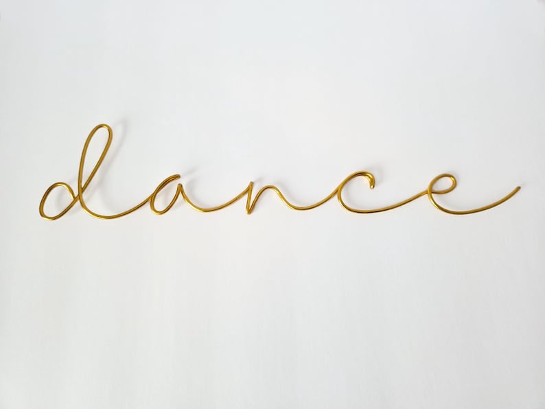 Op de afbeelding: Een goudkleurige draadsculptuur met het woord 'dance' in een cursief lettertype. Het woord is gecentreerd op een witte achtergrond. De draad is dun en heeft een gladde, glanzende afwerking, wat een minimalistische en elegante esthetiek cre&euml;ert.