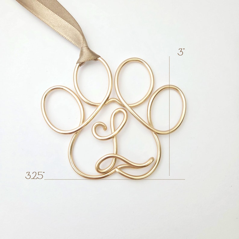 May include: A gold-colored wire paw print ornament with the letter "L" in the center. The ornament is 3.25 inches wide and 3 inches tall, with a tan ribbon for hanging. The paw print design is a decorative item.