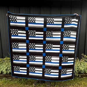 May include: A quilt featuring a grid of sixteen rectangular blocks, each displaying a stylized American flag pattern in black, white, and gray with a single horizontal blue stripe. The blocks are separated by black sashing and blue cornerstones.