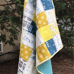 May include: A patchwork quilt with a yellow, blue, and white color scheme. The quilt is made of various fabrics with different patterns, including owls, flowers, and geometric designs. The quilt is folded and hanging from a tree branch.