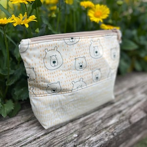 May include: A cream-colored zippered pouch with a repeating pattern of cartoon bear faces and small orange dots. The bottom section of the pouch has a light yellow vertical stripe pattern. The pouch is sitting on a wooden surface.