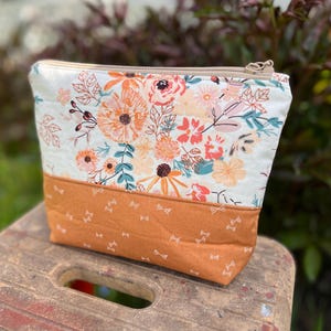 May include: A rectangular cosmetic bag with a floral pattern and a solid orange bottom. The top section features a cream background with orange, pink, and teal flowers. The bottom section is a solid orange fabric with small white bow accents.