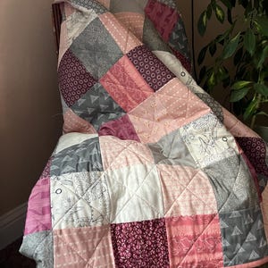 May include: A patchwork quilt draped over a chair. The quilt features squares of various pink, gray, and white fabrics with different patterns, including floral, geometric, and solid designs. The quilt has a pink border and is quilted with a diamond pattern.