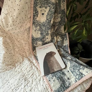 May include: A quilted blanket with a white background and black polka dots. The blanket has a patterned border with a dog design. A book titled "I Want My Hat Back" is on top of the blanket.