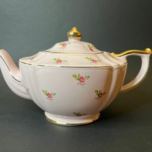 May include: A pink teapot with a gold trim and a floral pattern. The teapot has a lid with a gold knob and a spout on the side.