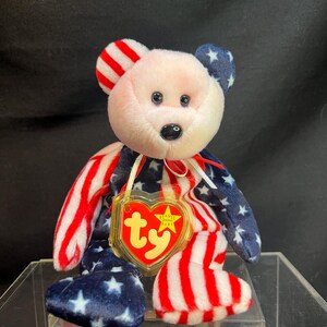 May include: A patriotic plush bear with a pink face, red and white striped arms and feet, and blue star-covered ears and legs. It has a heart-shaped tag with the text "Ty Beanie Baby".
