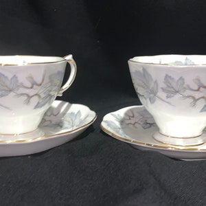 Puede incluir: Dos tazas de té blancas y platillos con un delicado diseño de hojas plateadas y grises. Las tazas tienen un borde dorado y los platillos tienen un borde festoneado.