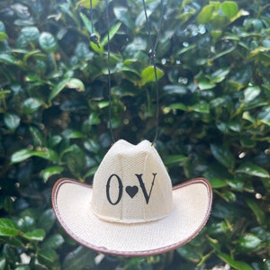 May include: A white cowboy hat with a brown brim, hanging from a black string. The hat has the letters "O" and "V" with a black heart in between.