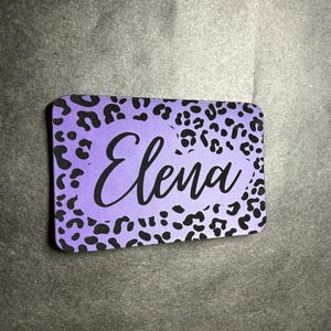 Puede incluir: Una etiqueta con nombre rectangular con esquinas redondeadas. La etiqueta presenta un fondo morado con un estampado de leopardo negro. El nombre "Elena" está escrito en escritura cursiva negra en el centro de la etiqueta.