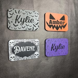 Puede incluir: Cuatro etiquetas rectangulares con esquinas redondeadas. Una es gris con murciélagos negros y el nombre Kylie. Una es naranja con una cara de calabaza negra y el nombre Amber. Una es negra con calaveras y rosas blancas y el nombre DAVENE. Una es morada con fantasmas blancos y el nombre Kylie.
