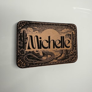 May include: A rectangular, brown wooden sign with the name "Michelle" in black, featuring a cowboy theme. The design includes a cowboy hat, boots, rope, cacti, and a sunset. The border is decorated with floral and rope details.