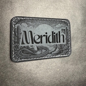 May include: Rectangular black leather patch with white stitching. The patch features a desert landscape design with the name "Meredith" in large letters. The design includes a cowboy hat, boots, rope, and cacti.