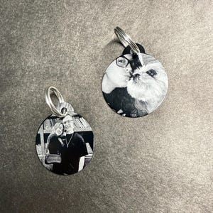 May include: Two round, black and white metal keychains. One keychain features a couple, the other a person kissing a dog. Each keychain has a silver ring for attachment. The background is a dark gray surface.