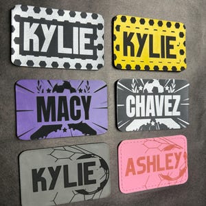 May include: Six rectangular name tags with soccer-themed designs. The tags feature names like "KYLIE," "MACY," "CHAVEZ," and "ASHLEY" in various colors, including black, yellow, purple, gray, and pink. Each tag has a soccer ball or soccer shoe graphic.
