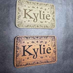 May include: Two rectangular patches with rounded corners, each featuring a paisley border pattern and the name Kylie centered in a serif font. One patch is light beige with dark brown details, while the other is tan with black details.