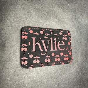 May include: A rectangular black patch with a pink cherry pattern and the name "Kylie" in pink script. The patch has rounded corners and a stitched edge, set against a gray background.