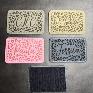 Może przedstawiać: Kolekcja spersonalizowanych naszywek z imionami z wzorem w panterkę. Naszywki są prostokątne z zaokrąglonymi rogami i zawierają imiona lub inicjały w różnych czcionkach. Kolory obejmują szary, różowy i niebieski. Widoczne jest również czarne zapięcie na rzep.