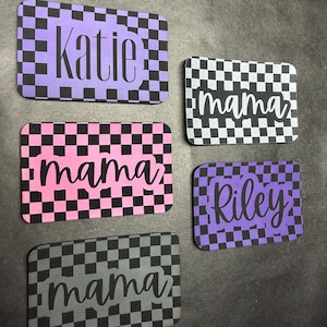 May include: Rectangular name tags with a checkered pattern in purple, pink, black, and gray. The tags feature names like "Katie" and "Riley," and the word "mama" in cursive. The background is a dark gray surface.