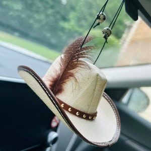 May include: A white cowboy hat with a brown leather band and gold studs hanging from a rearview mirror in a car. A brown and white feather is attached to the hat.