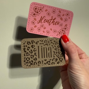 Może przedstawiać: Dwie prostokątne skórzane naszywki. Górna naszywka jest różowa z imieniem "Heather" w kursywie i kwiatowymi akcentami. Dolna naszywka jest beżowa z nadrukiem w cętki i imieniem "Tina" wielkimi literami.