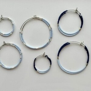 May include: Six pairs of hoop earrings in different sizes. The earrings feature a silver metal base and are decorated with small, light blue and navy blue beads. They are arranged on a white surface.