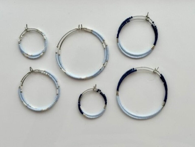 May include: Six pairs of hoop earrings in different sizes. The earrings feature a silver metal base and are decorated with small, light blue and navy blue beads. They are arranged on a white surface.