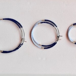 May include: Three pairs of hoop earrings in different sizes. Each earring has a silver-coloured metal hoop with a double row of small, light blue and navy blue beads. The earrings are displayed on a white surface.