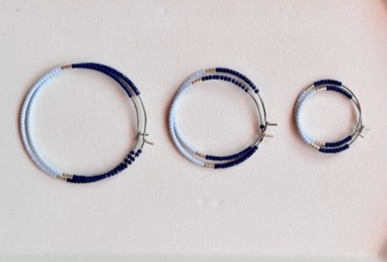 May include: Three pairs of hoop earrings in different sizes. Each earring has a silver-coloured metal hoop with a double row of small, light blue and navy blue beads. The earrings are displayed on a white surface.