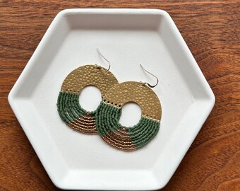 May include: A pair of gold-toned, hammered metal hoop earrings with a circular design. The lower half of each earring is adorned with rows of green and bronze-colored beads. The earrings are displayed on a white, hexagonal tray, set against a wooden background.