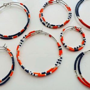 May include: Several pairs of hoop earrings with a beaded design. The earrings feature a combination of navy blue, orange, white, and silver beads. The hoops are made of silver-colored metal and are displayed on a white surface.