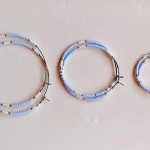 May include: Three beaded hoop earrings in different sizes. The earrings are made with light blue and white beads on a wire frame. They are displayed on a pale pink surface.