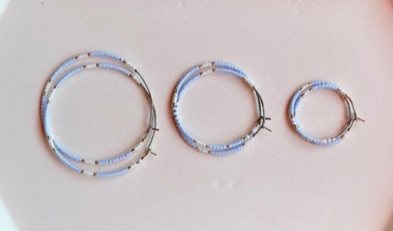 May include: Three beaded hoop earrings in different sizes. The earrings are made with light blue and white beads on a wire frame. They are displayed on a pale pink surface.