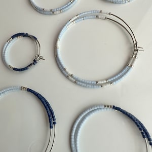May include: A collection of beaded hoop earrings in different sizes. The earrings are adorned with light blue, white, and navy blue beads. The metal hoops are silver. The earrings are presented on a white surface.
