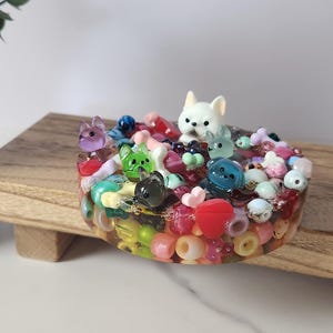 K&ouml;nnte beinhalten: Ein farbenfrohes Kunstwerk aus Kunstharz mit kleinen Tierfiguren, Perlen und Herzformen in verschiedenen Farben. Das Kunstwerk befindet sich auf einer Holzoberfl&auml;che. Es ist ein dekoratives Objekt.