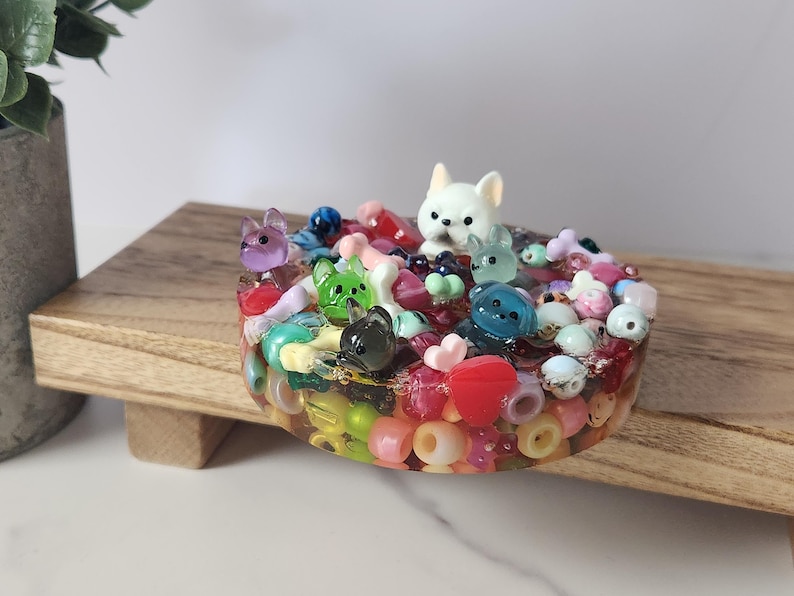K&ouml;nnte beinhalten: Ein farbenfrohes Kunstwerk aus Kunstharz mit kleinen Tierfiguren, Perlen und Herzformen in verschiedenen Farben. Das Kunstwerk befindet sich auf einer Holzoberfl&auml;che. Es ist ein dekoratives Objekt.