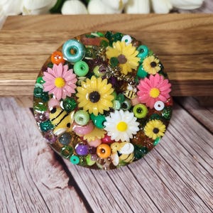 May include: A round, decorative resin piece with a floral and bee theme. The design features yellow sunflowers, pink daisies, and white daisies, along with green beads, and small bee figurines. The piece is set on a wooden surface.
