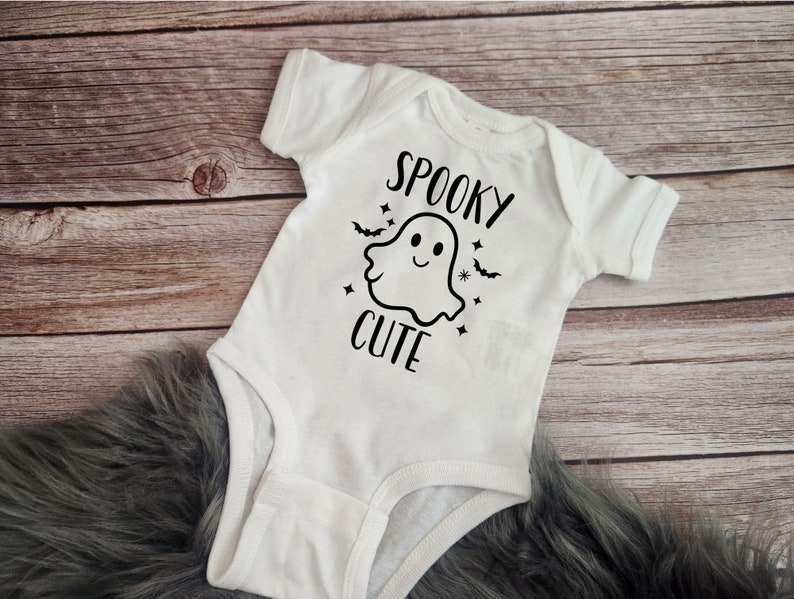 Pode incluir: Macac&atilde;o branco para beb&ecirc; com um gr&aacute;fico de fantasma preto e o texto "Spooky Cute".