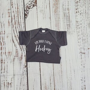 May include: A gray baby onesie with the text "I'M TOLD I LOVE Hockey" in white script. The onesie is laid flat on a weathered, light-colored wooden surface.