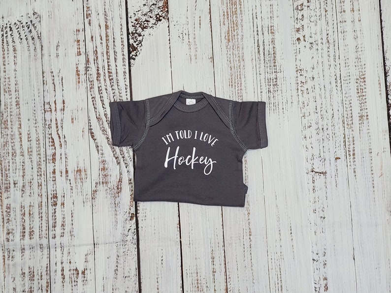 May include: A gray baby onesie with the text "I'M TOLD I LOVE Hockey" in white script. The onesie is laid flat on a weathered, light-colored wooden surface.