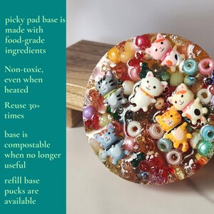 K&ouml;nnte beinhalten: Eine runde, transparente Harzbasis mit bunten Perlen und kleinen Katzenfiguren. Die Basis ist mit verschiedenen Farben von Perlen und kleinen Katzenfiguren dekoriert. Der Text auf der linken Seite lautet "picky pad base is made with food-grade ingredients."