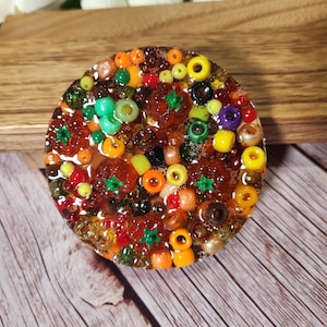 May include: A round, colorful resin coaster with embedded beads. The coaster features a mix of orange, green, yellow, red, and brown beads, creating a textured, mosaic-like appearance. The coaster is set against a wooden background.