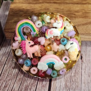 May include: A round, resin-based decorative piece featuring a whimsical design. It includes a pink unicorn, a rainbow, and various colorful beads and charms. The piece is approximately 3 inches in diameter.
